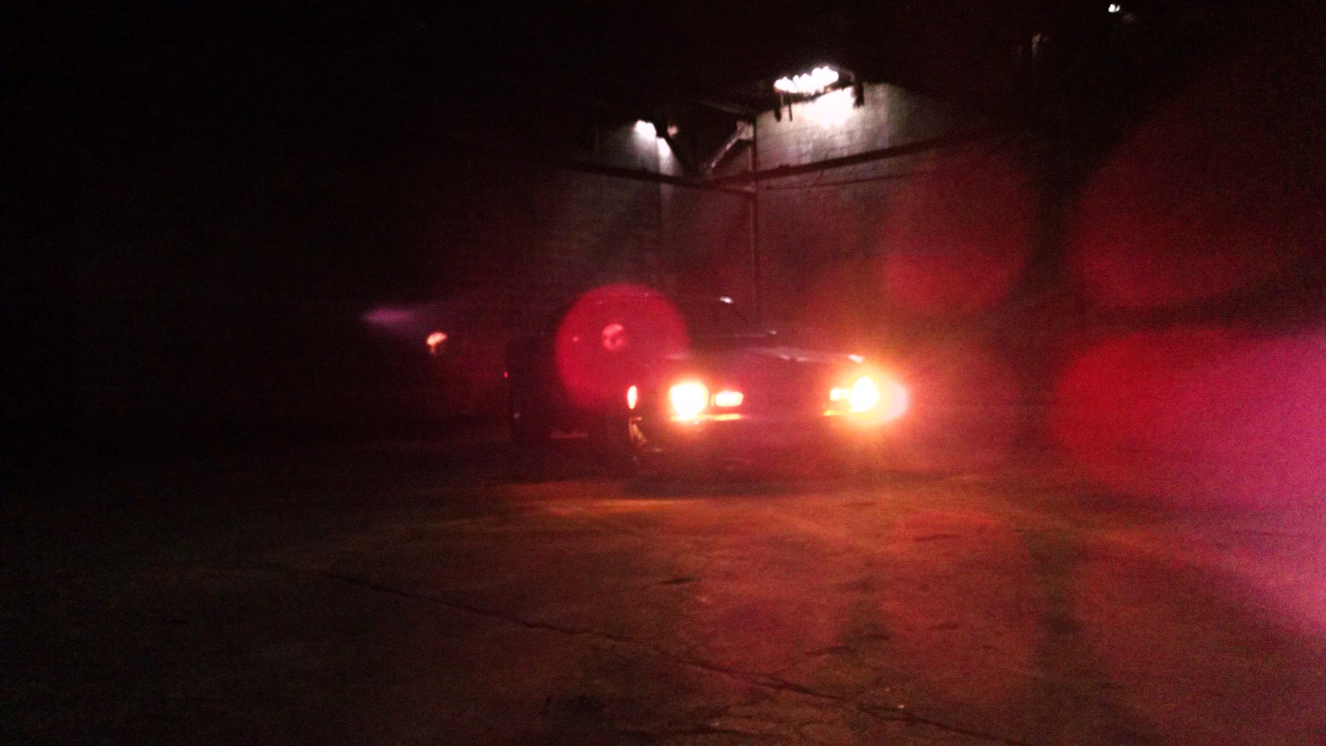 Muscle car with headlights on, lit red inside a dark garage